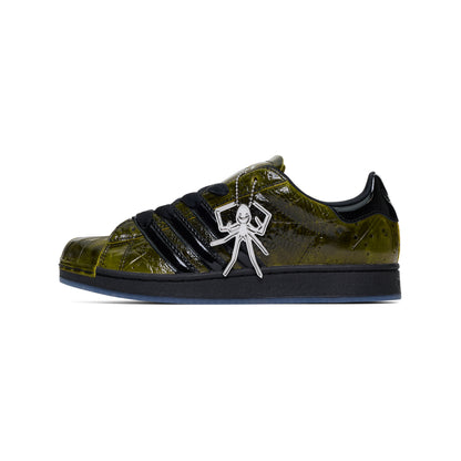 SP5DER x ADIDAS SUPERSTAR sneaker with glossy cracked olive patent leather, black stripes, and a silver spider charm.