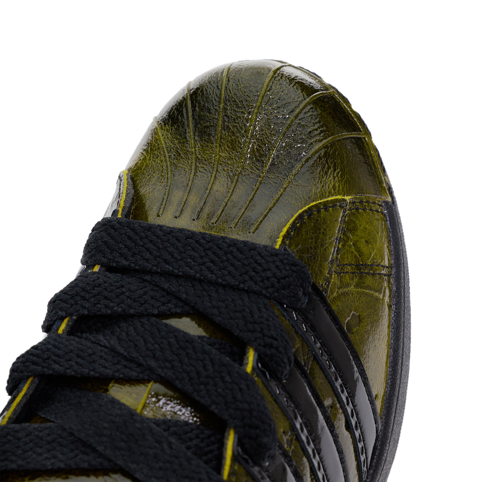 SP5DER x ADIDAS SUPERSTAR sneaker with glossy cracked olive patent leather, black laces, and signature serrated stripes.