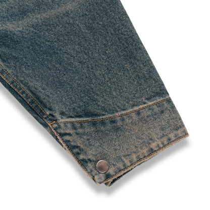 Worn blue denim work jacket sleeve with a single silver snap button cuff and contrast stitching.