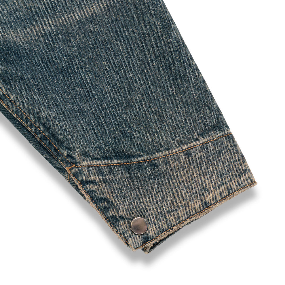 Worn blue denim work jacket sleeve with a single silver snap button cuff and contrast stitching.
