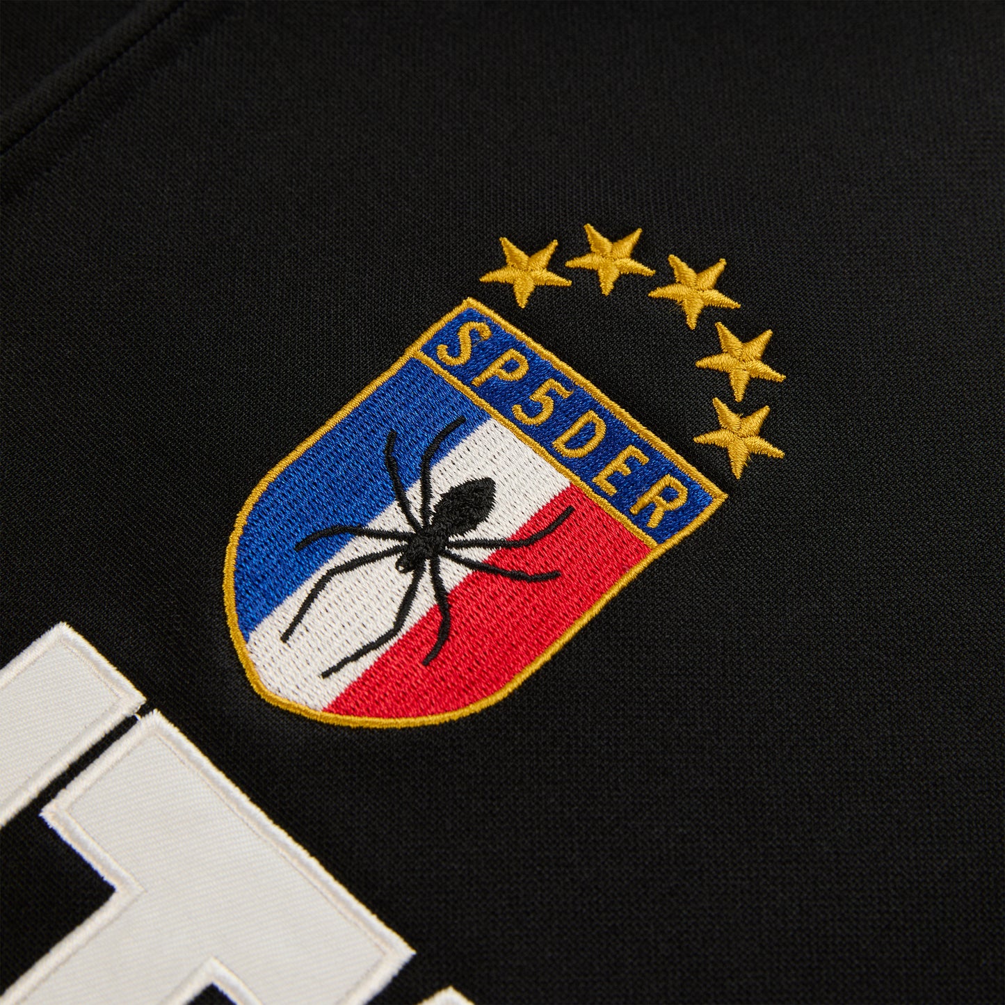 Close-up of SP5DER branded crest logo patch with spider and stars on black Atlanta Track Jacket.