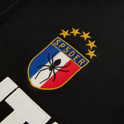 Close-up of SP5DER branded crest logo patch with spider and stars on black Atlanta Track Jacket.