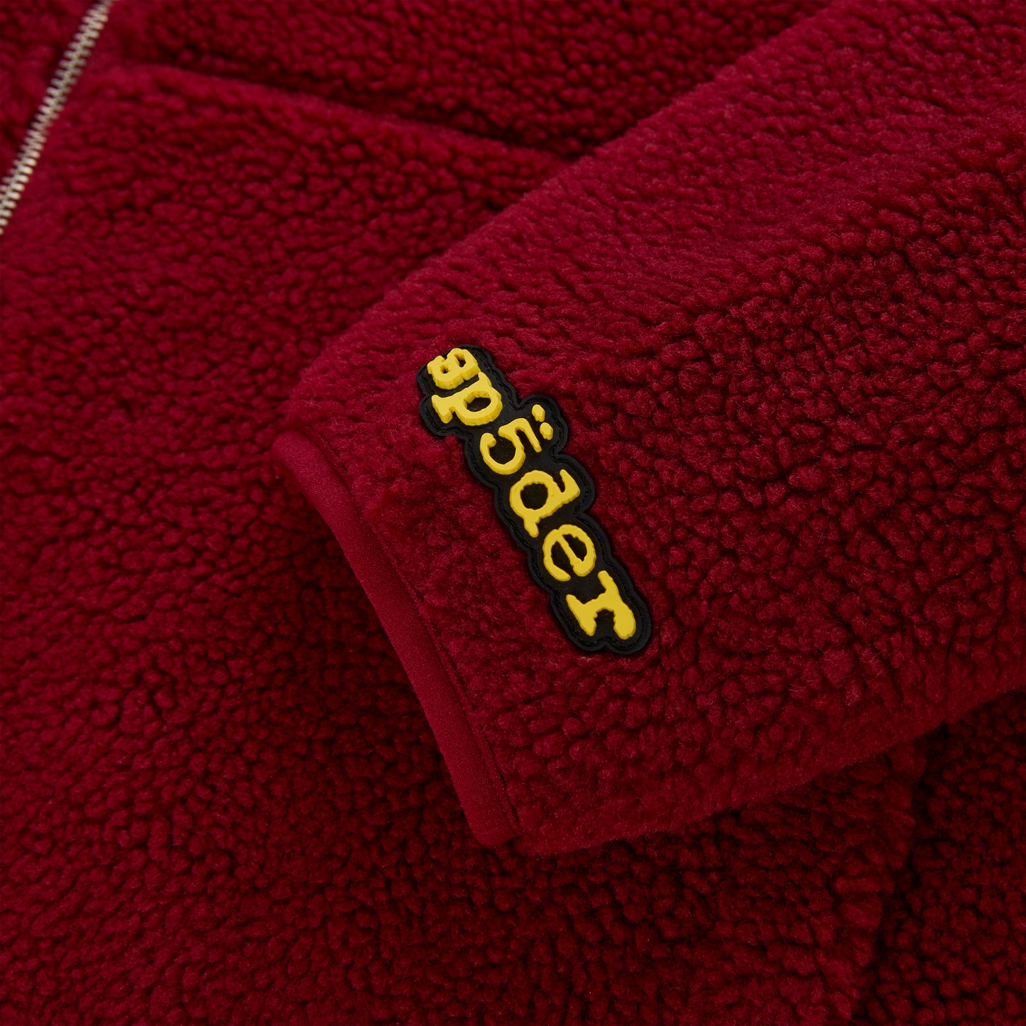 Close-up of a red sherpa fleece zip-up jacket sleeve with a yellow rubberized "Spyder" logo patch.