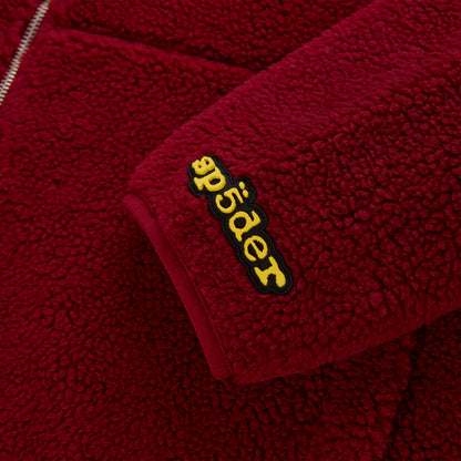 Close-up of a red sherpa fleece zip-up jacket sleeve with a yellow rubberized "Spyder" logo patch.