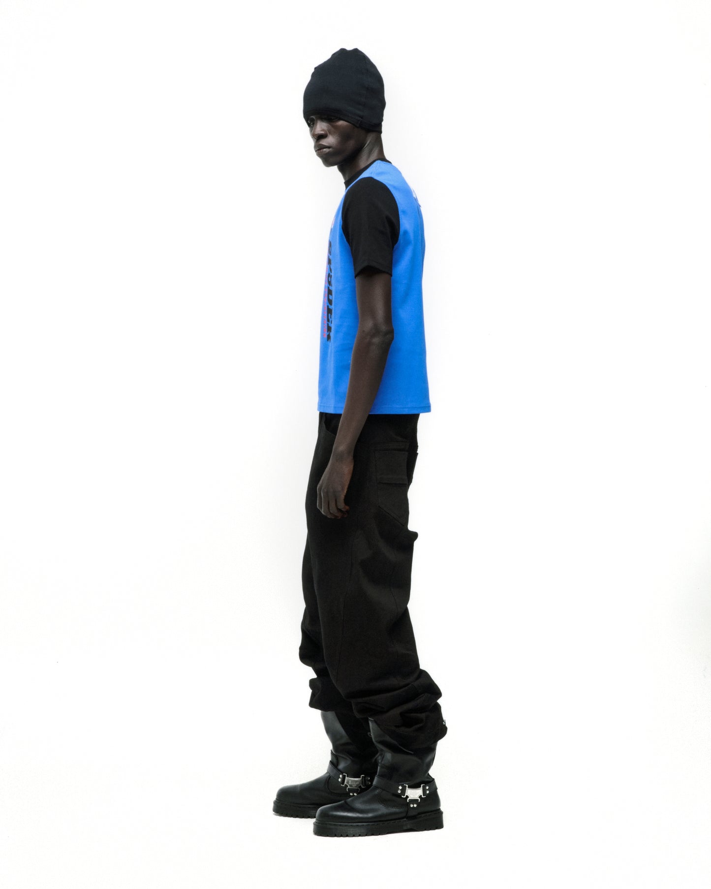 Black model wearing a black beanie, black tee, blue vest with graphic, black baggy pants, and black boots.