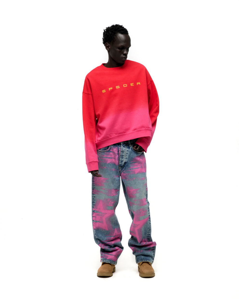 Model wears a red to pink gradient crewneck with yellow SP5DER text and star-patterned jeans.