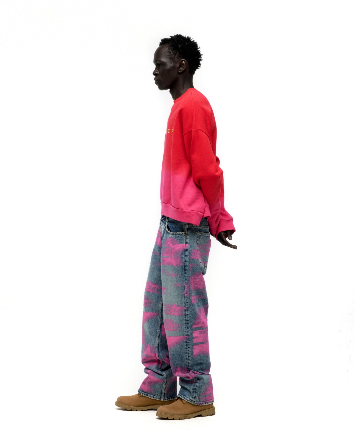 Man in red to pink gradient crewneck with SP5DER text, blue jeans with pink graphics, and brown boots.