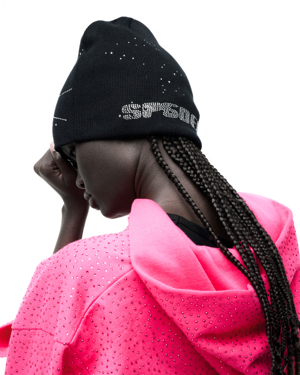 Black acrylic beanie with rhinestone skull design, worn by model with braids and a pink embellished hoodie.