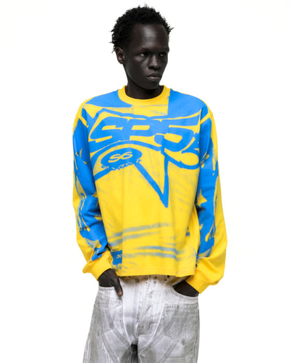Model wearing a yellow SP5 Winners long sleeve tee with blue graphic print, ribbed collar, and cuffs.