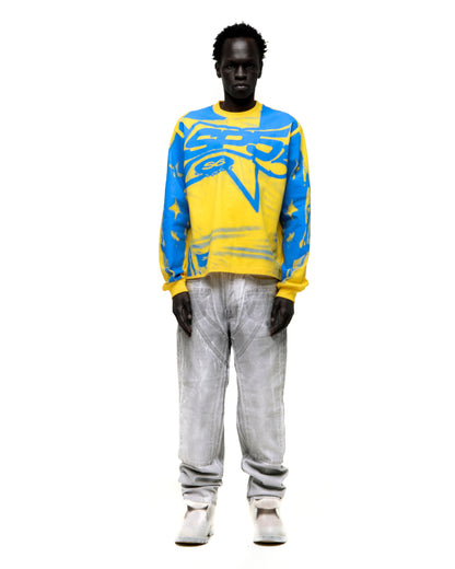 Model wearing yellow and blue SP5 WINNERS LONGSLEEVE with graphic print, ribbed collar/cuffs, and light grey jeans.