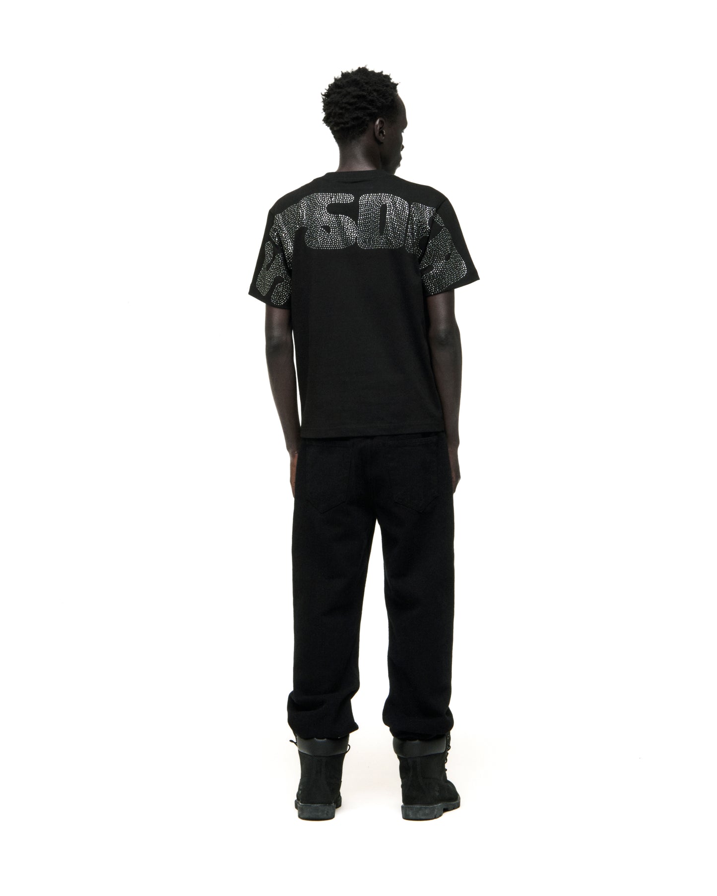 Man in black cotton rhinestone embellished tee with graphic on back, paired with black pants and boots.