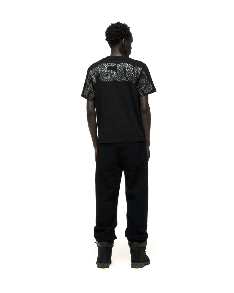 Man in black cotton rhinestone embellished tee with graphic on back, paired with black pants and boots.