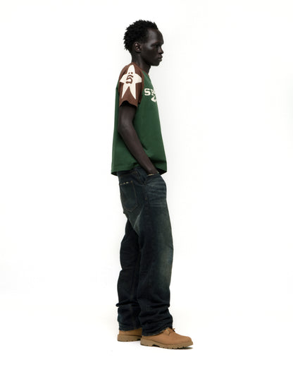 Man in 5UKI TEE: green cotton tee with brown color-block sleeves, graphic print, and dark jeans.