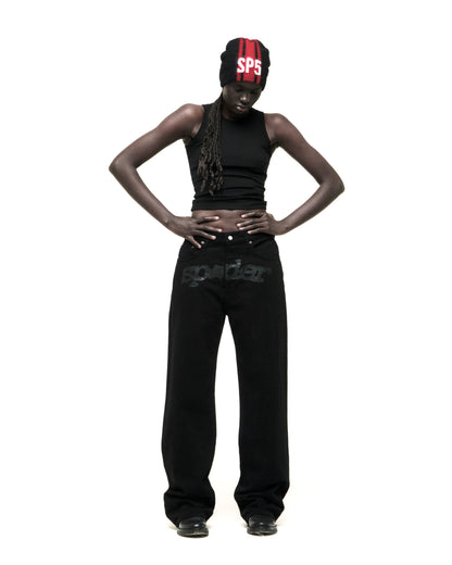 Model wearing black V3 DENIM pants with "spider" logo at crotch, black tank top, and red/black beanie.