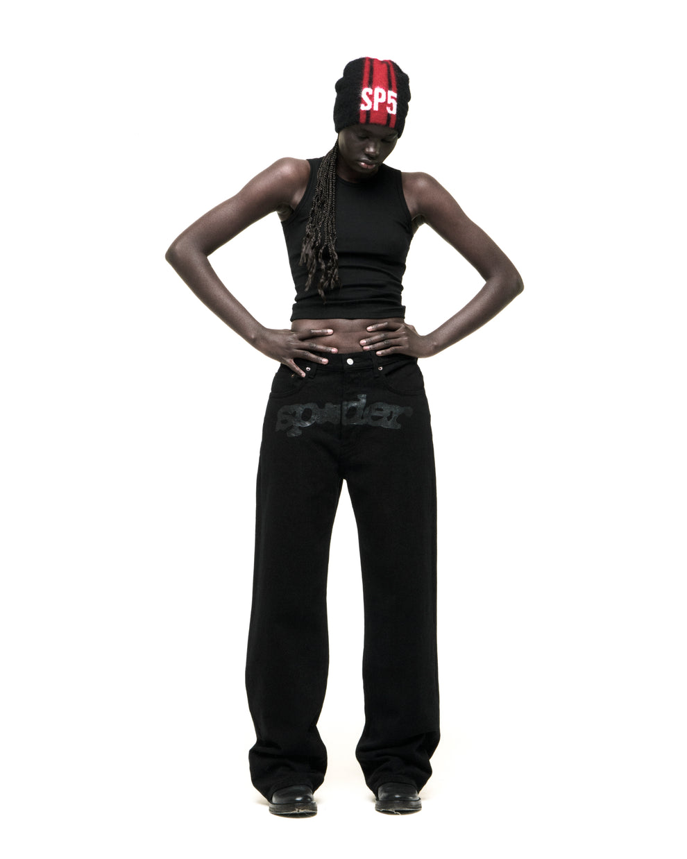 Model wearing black V3 DENIM pants with "spider" logo at crotch, black tank top, and red/black beanie.