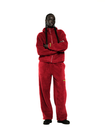 Model in a red sherpa fleece zip-up hooded jacket and matching pants with yellow logo patches.