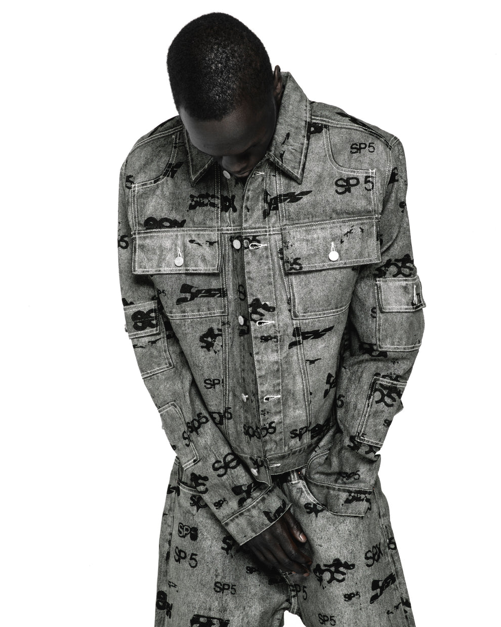 Gray denim trucker jacket with all-over black "SP5" print, silver buttons, and red contrast stitching.