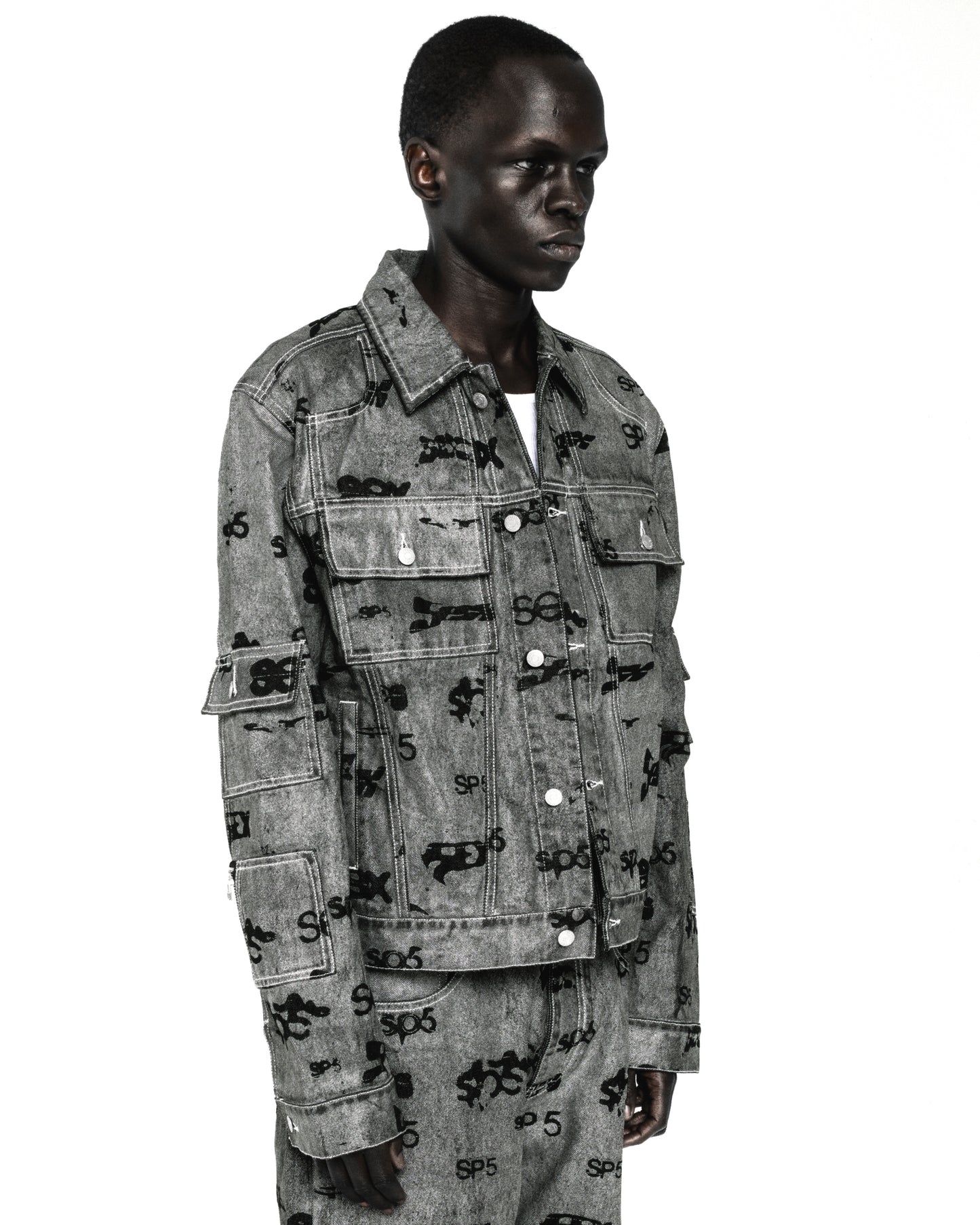 Black model wearing a gray denim trucker jacket with black all-over print, silver buttons, and arm pockets.