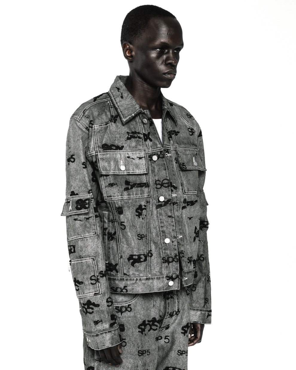 Black model wearing a gray denim trucker jacket with black all-over print, silver buttons, and arm pockets.