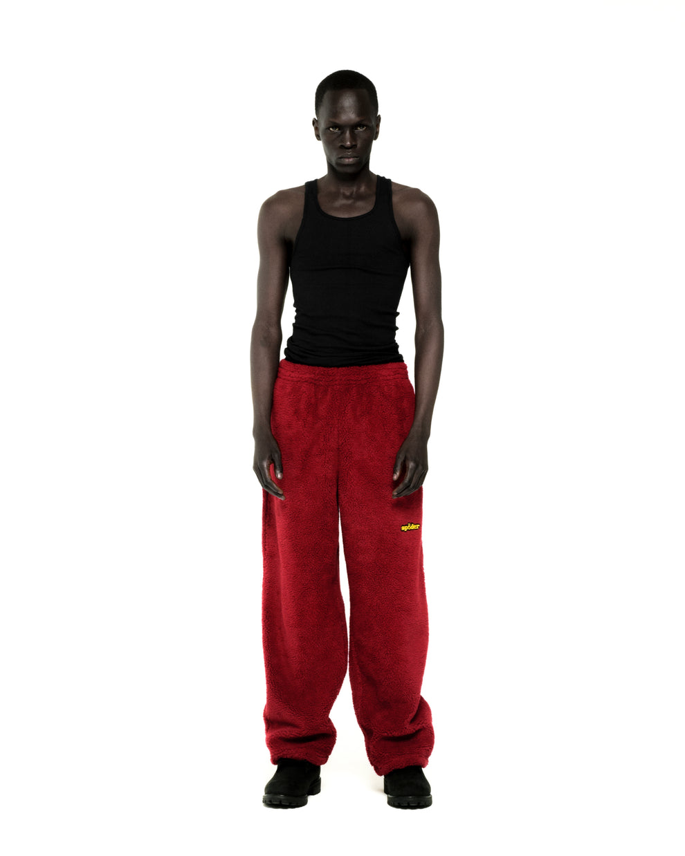 Model wearing red teddy fleece pants with yellow logo patch and black tank top.