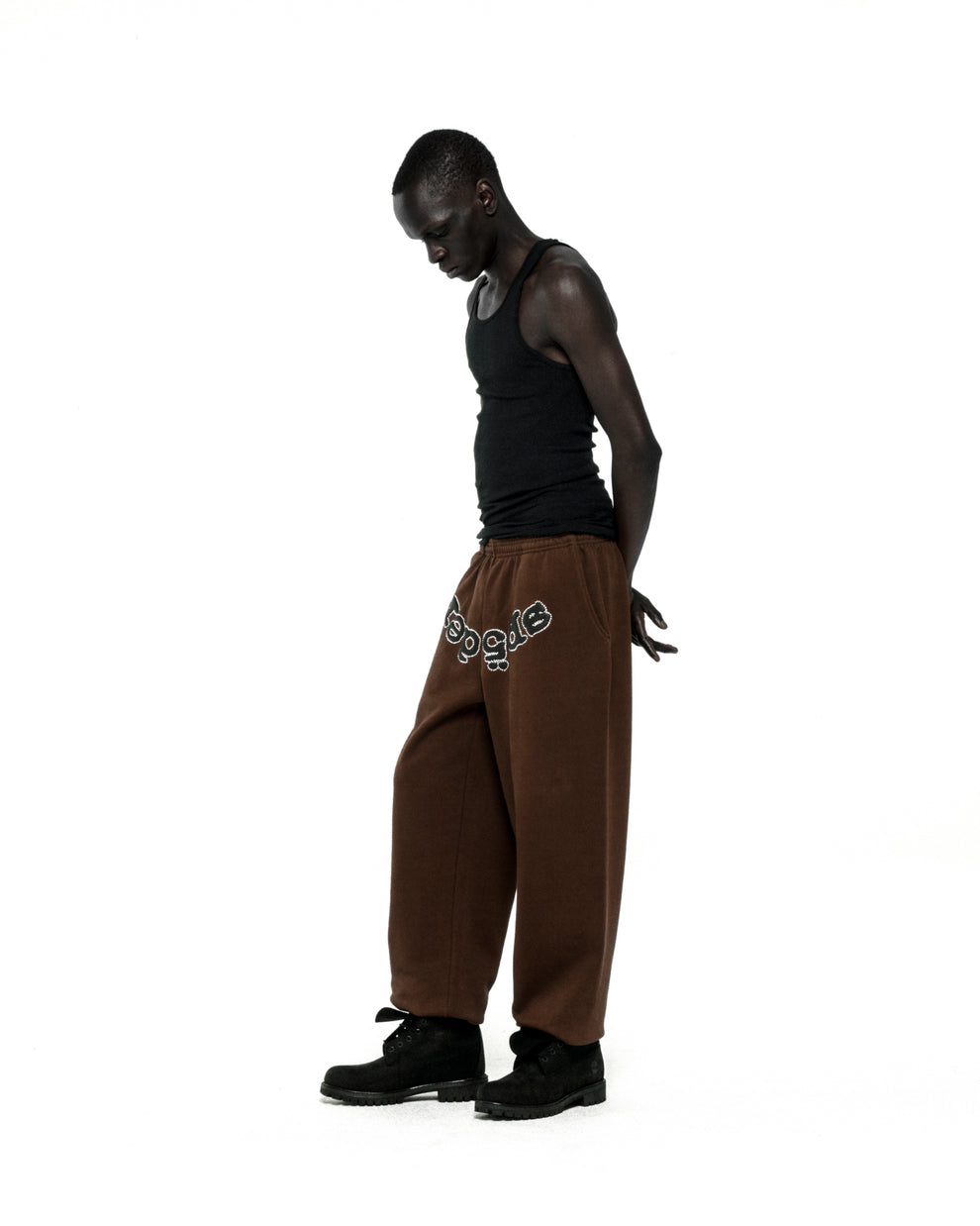 Brown cotton fleece sweatpants with "SP5DER" rhinestone text on the crotch, worn by a model.