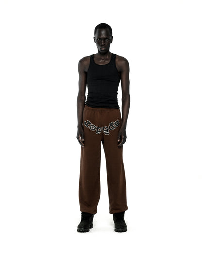 Brown fleece sweatpants with "SP5DER" rhinestone text on the crotch, worn by a model.