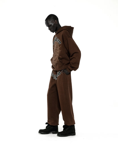 Model wearing a brown RHINESTONE OG WEB HOODIE and matching sweatpants with rhinestone spiderweb detail.