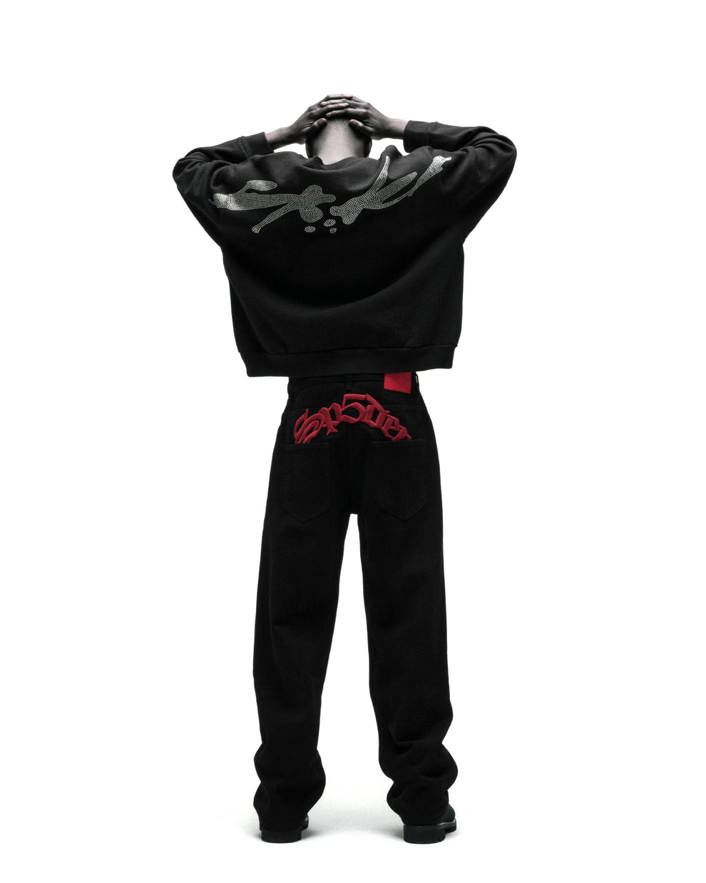 Black cotton crewneck with rhinestone graphic on back, paired with black jeans featuring red embroidered text.