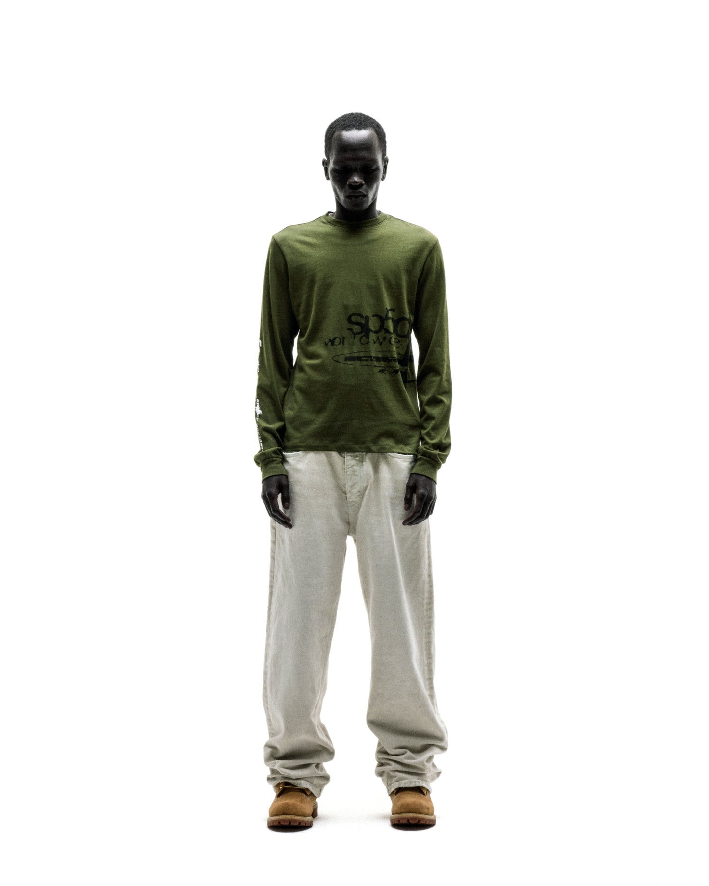 Dark-skinned model in an olive green cotton long sleeve tee with black graphic print, light linen pants, and brown boots.