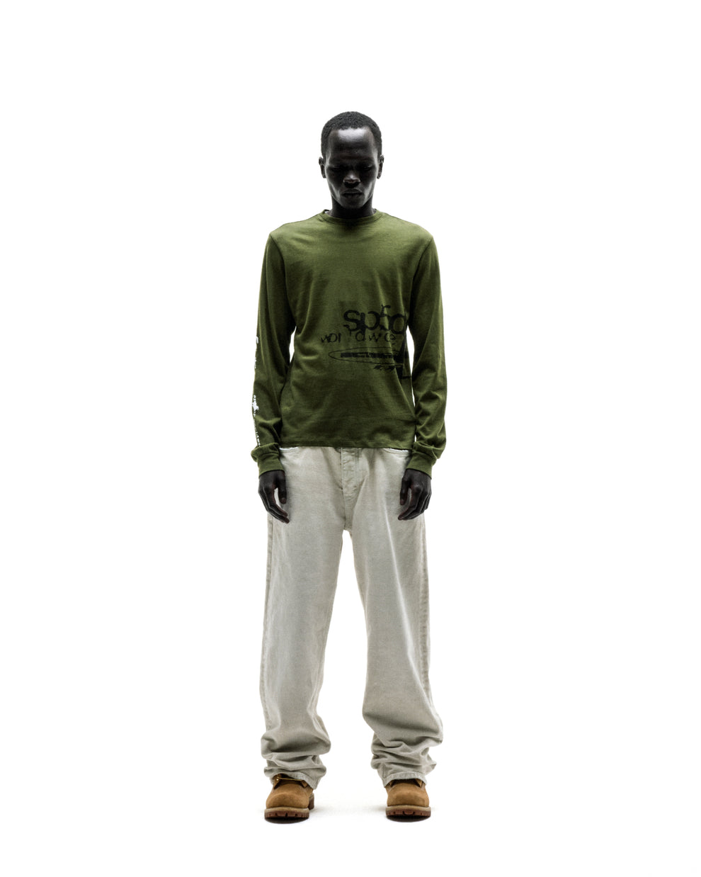 Dark-skinned model in an olive green cotton long sleeve tee with black graphic print, light linen pants, and brown boots.