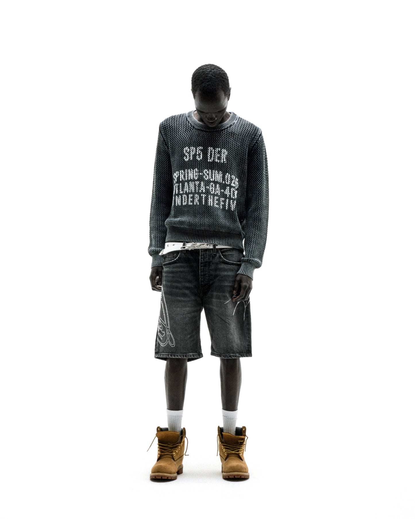 Black knit cotton UNDERTHEFIVE sweater with white text print, worn with dark denim shorts and tan boots.