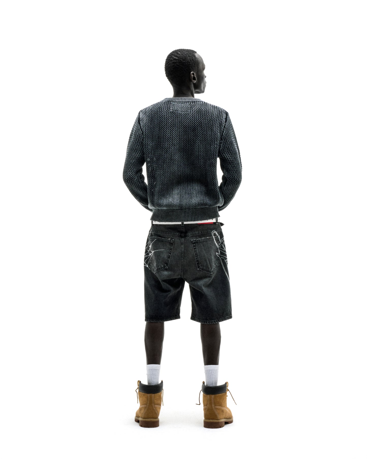 Man in dark knit cotton UNDERTHEFIVE sweater, black denim shorts with white graphics, and brown boots.