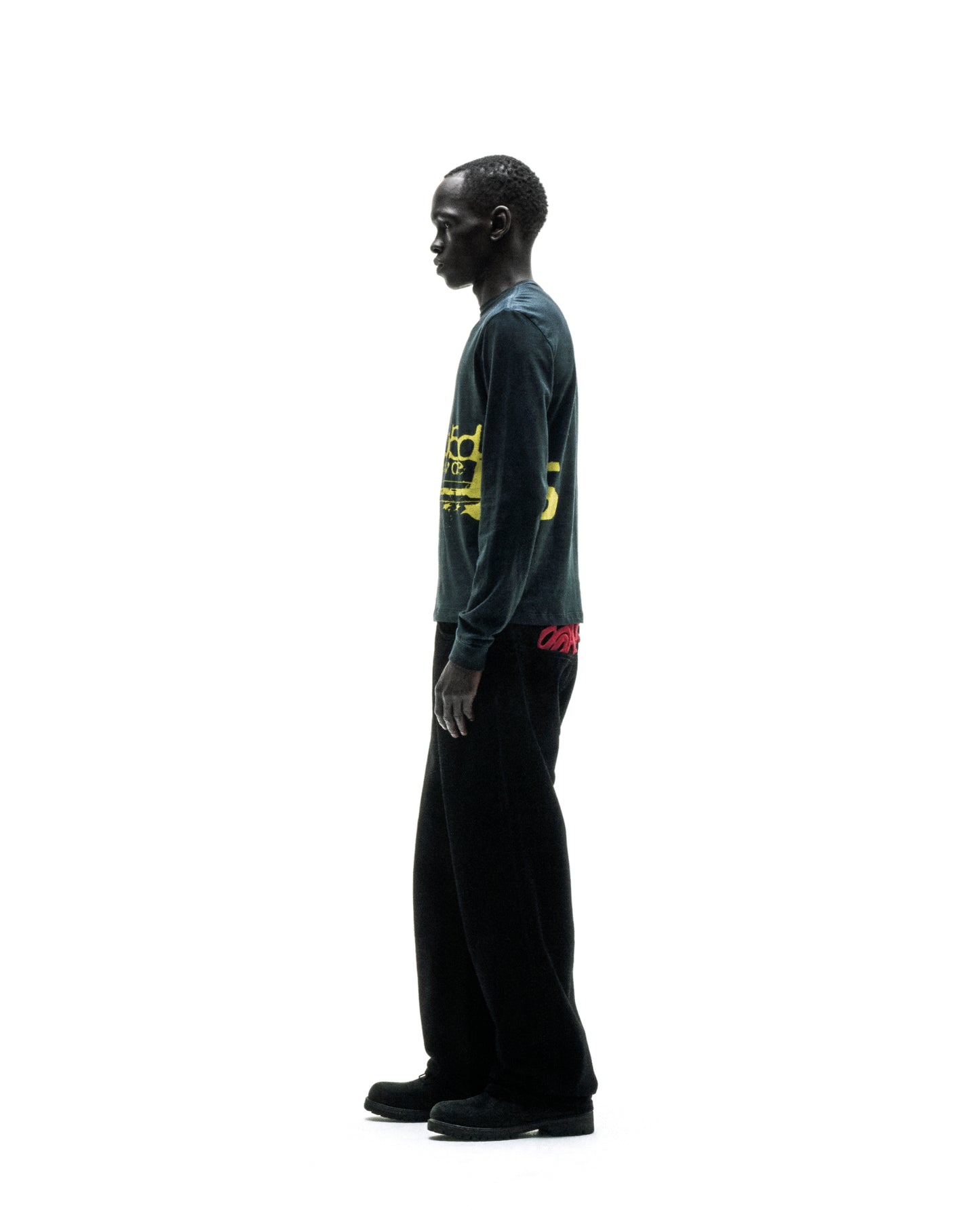 Man in dark green PURE ENERGY LONGSLEEVE with yellow graphic print, black pants, and shoes.
