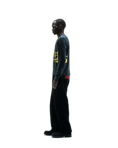 Man in dark green PURE ENERGY LONGSLEEVE with yellow graphic print, black pants, and shoes.