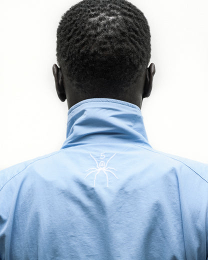 Back of a person wearing a light blue nylon track jacket with a white embroidered spider logo on the back neck.