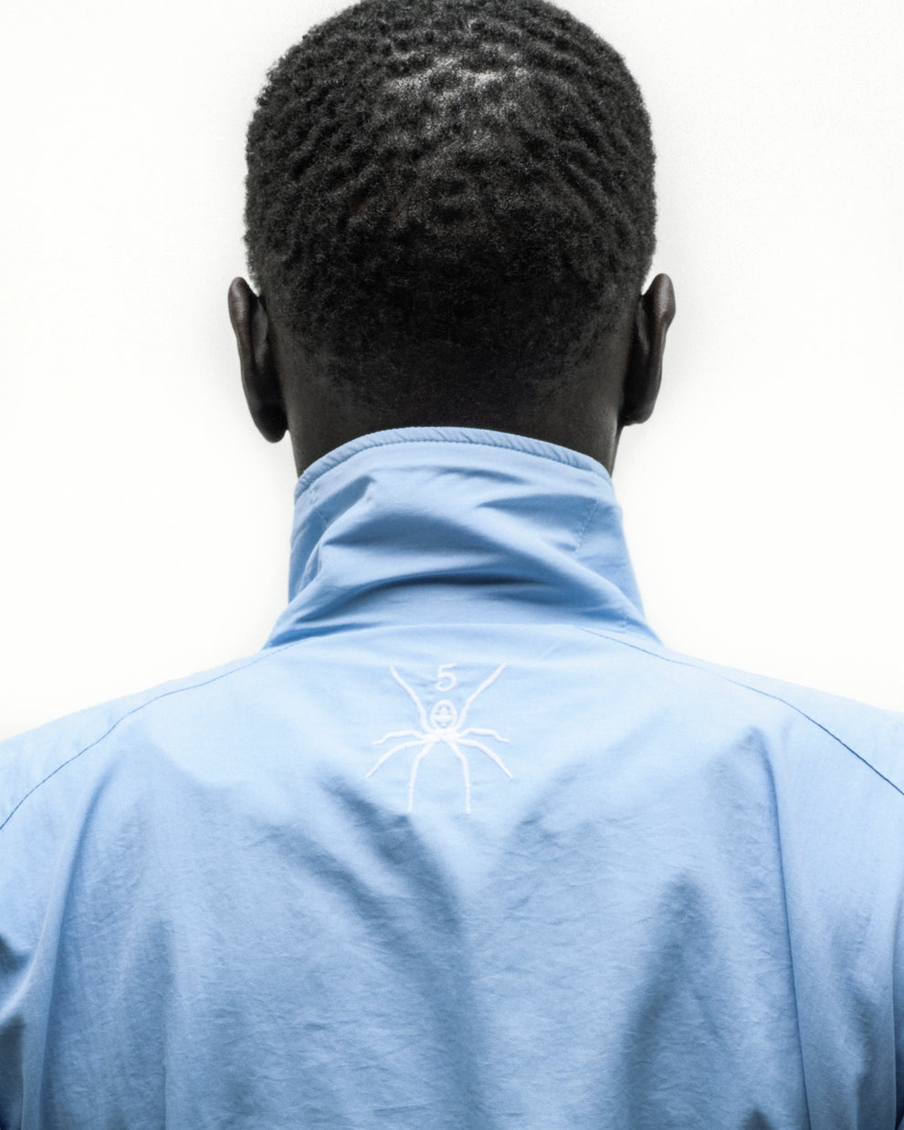 Back of a person wearing a light blue nylon track jacket with a white embroidered spider logo on the back neck.