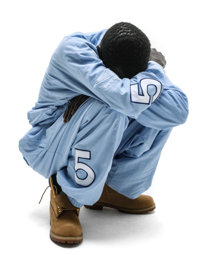 Man in light blue nylon track jacket and pants with "5" graphics, brown boots.