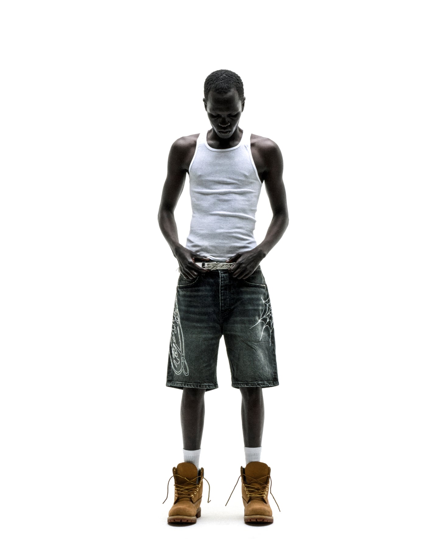 Man in vintage wash denim shorts with rhinestone web design, white tank top, and brown boots.