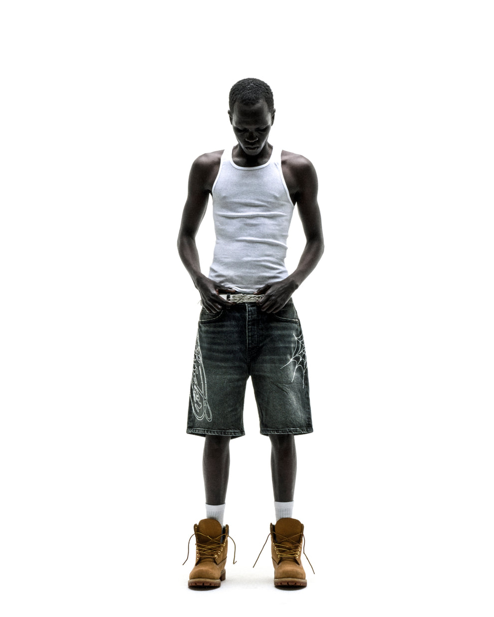 Man in vintage wash denim shorts with rhinestone web design, white tank top, and brown boots.
