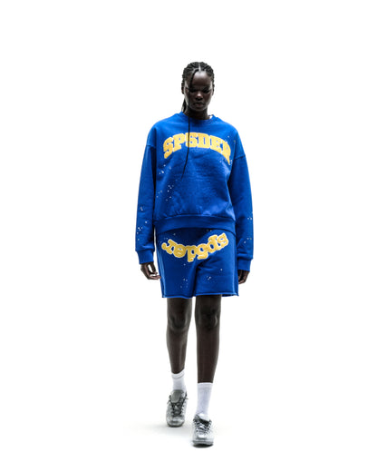 Blue crewneck sweatshirt with yellow varsity-style "SP5DER" text across the chest, ribbed cuffs, and relaxed fit.