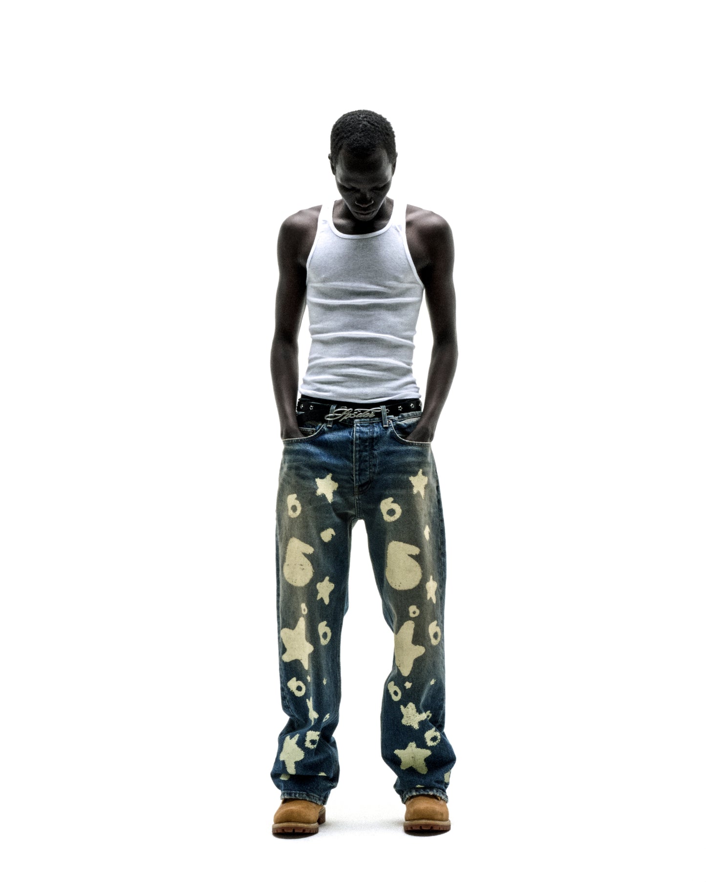 Man wearing Beluga Stamp Denim jeans with star and number designs, a white tank top, and tan boots.