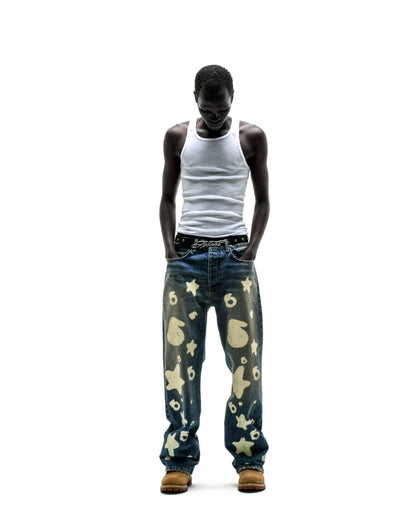 Man wearing Beluga Stamp Denim jeans with star and number designs, a white tank top, and tan boots.