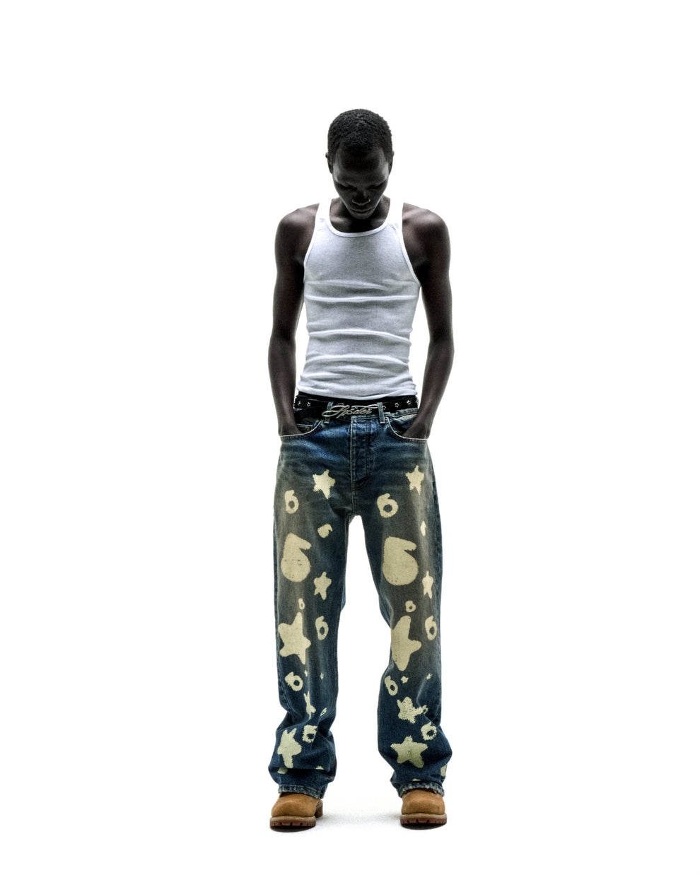 Man wearing Beluga Stamp Denim jeans with star and number designs, a white tank top, and tan boots.