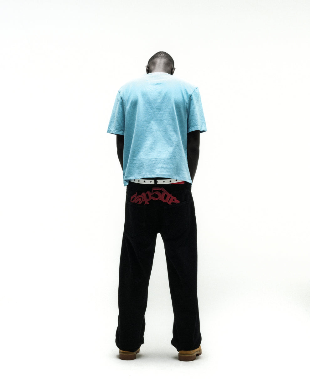 Man from behind wearing a light blue cotton tee and black pants with red embroidered text.