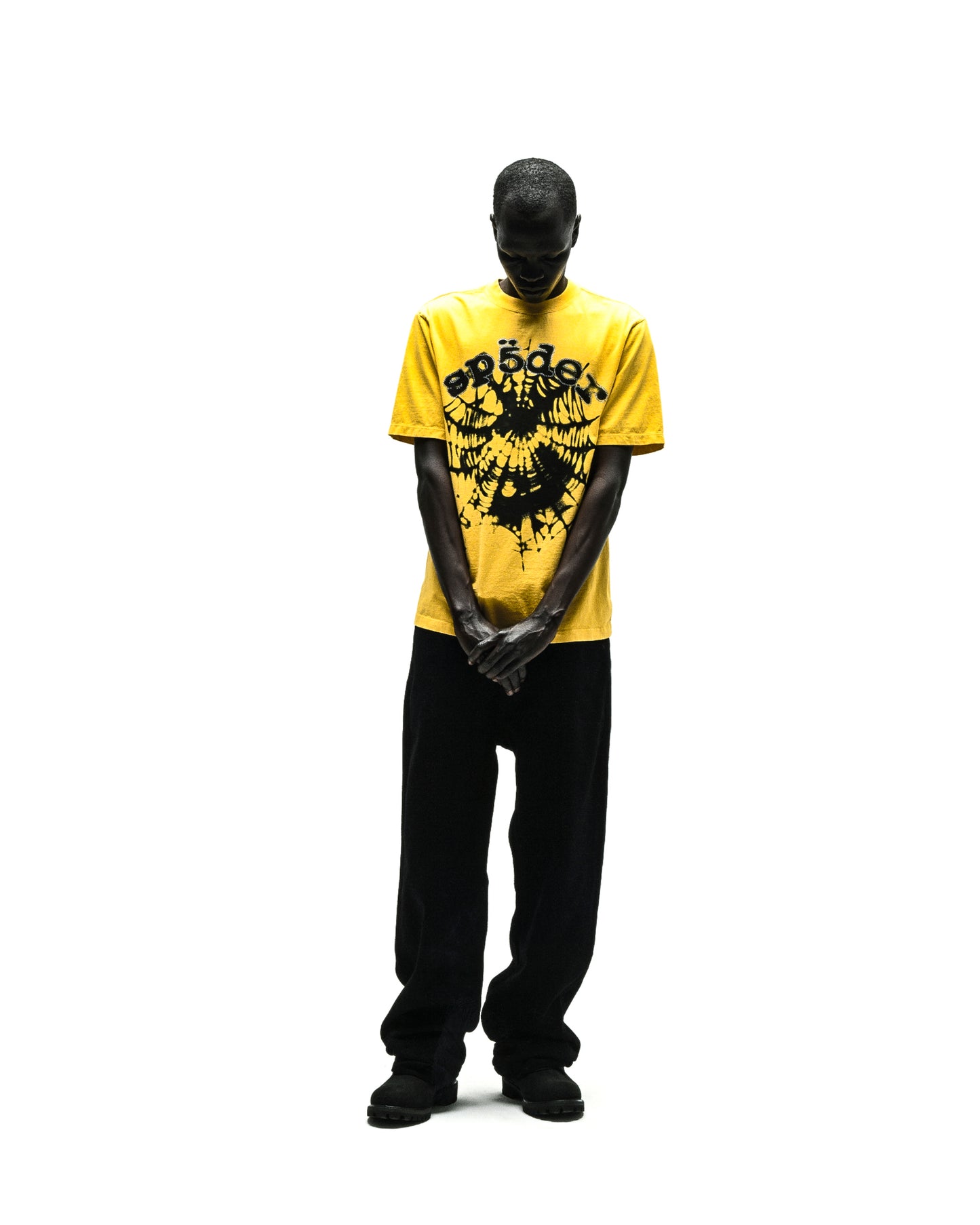 Man wearing a yellow SCATTERED OG WEB V2 TEE with black "SP5DER" text and rhinestone print, paired with black pants.