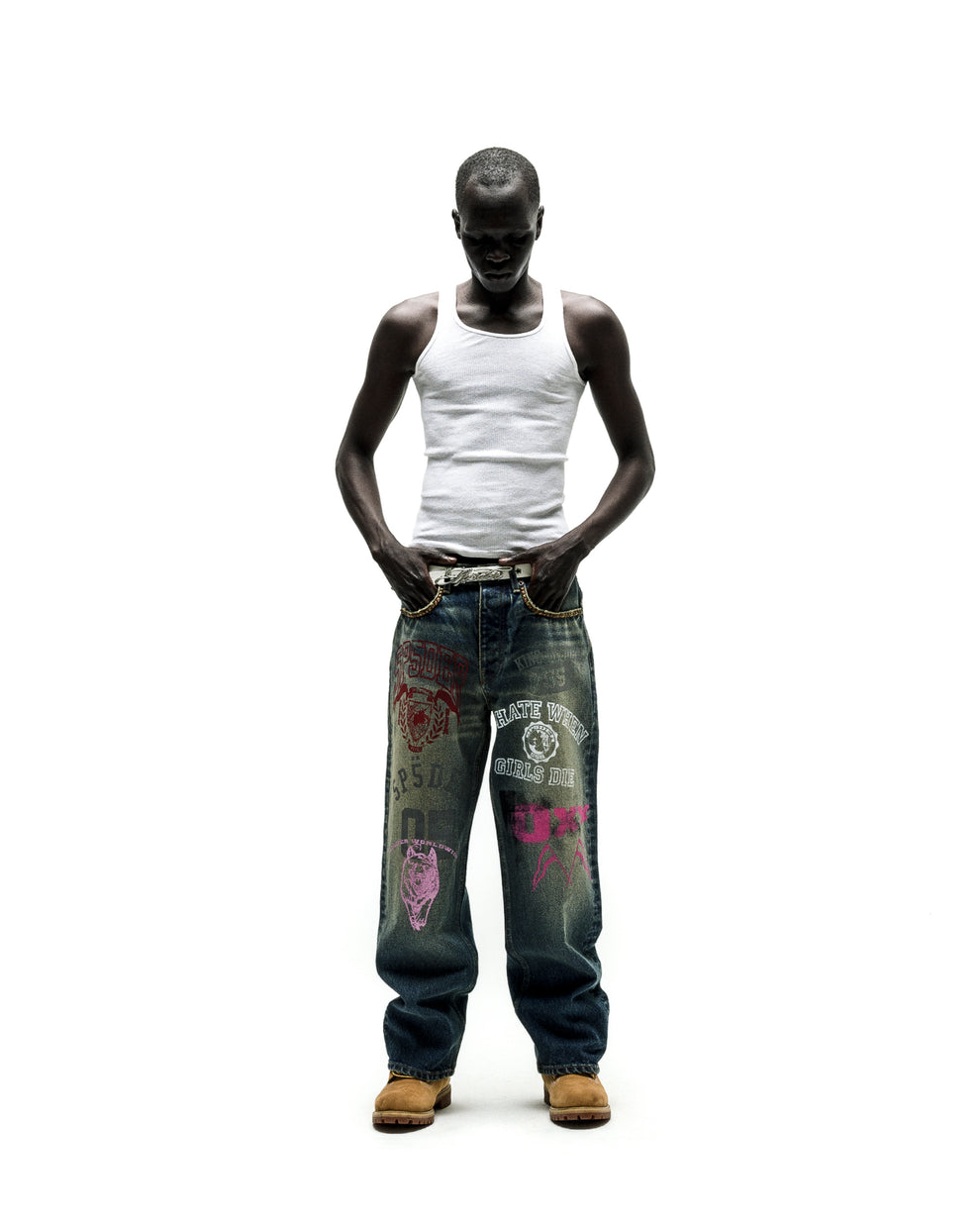 Man in a white tank top, baggy blue jeans with colorful distressed patches, and tan boots.