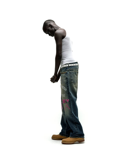 Man in white tank top, blue baggy jeans with pink graffiti print, and tan work boots.