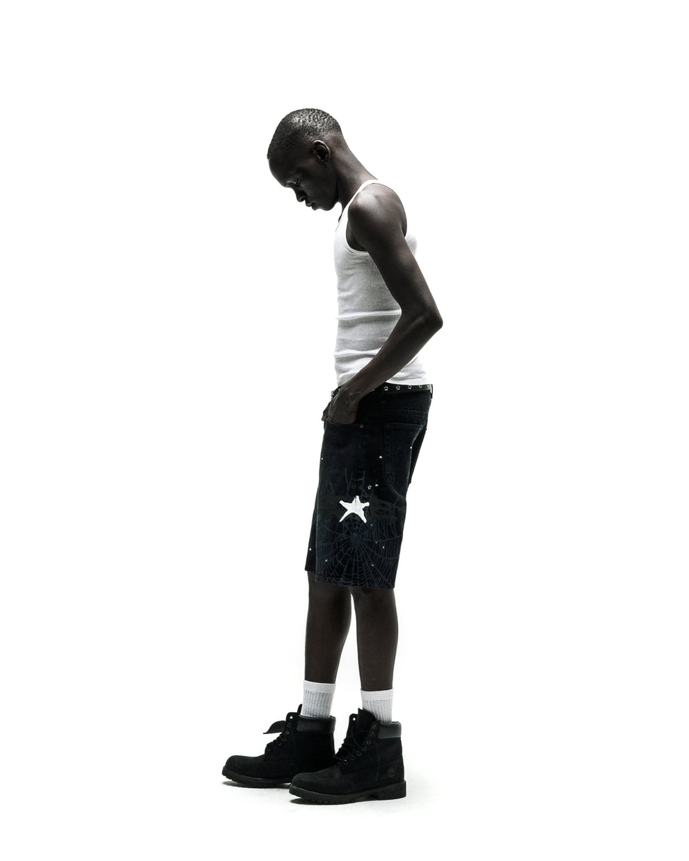 Man in dark wash HEAVY WEB JEAN SHORTS with white star graphic, white tank top, and black boots.