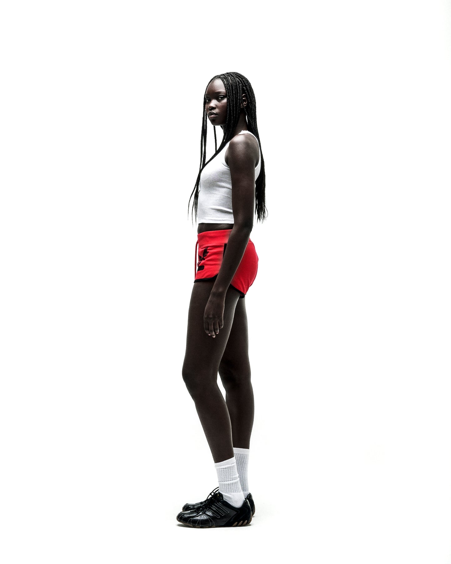 Woman in SP5 WIDE BOXER red shorts with black logo, white tank top, and black sneakers.