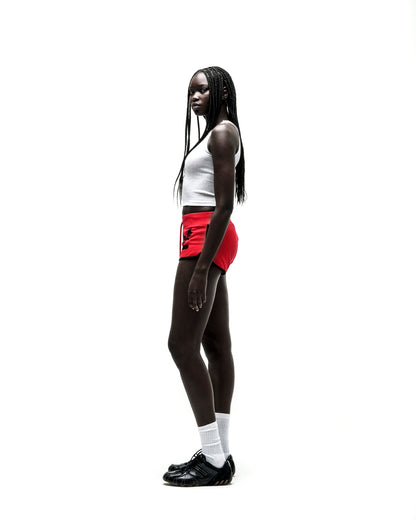 Woman in SP5 WIDE BOXER red shorts with black logo, white tank top, and black sneakers.